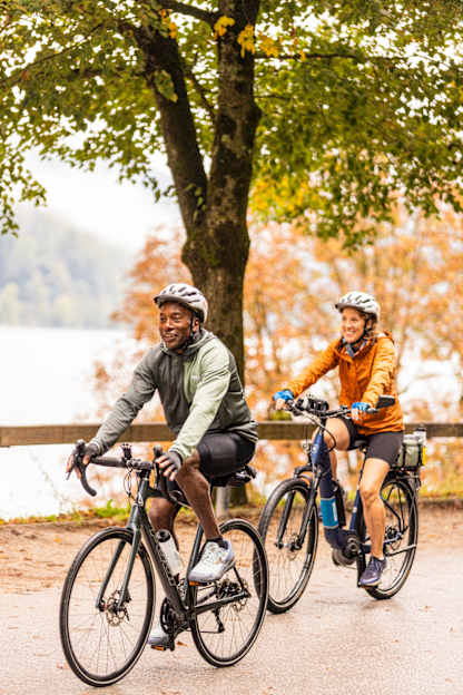 Two cyclists ride along a tree-lined path in autumn, smiling as they pedal past colorful foliage and a lakeside backdrop.