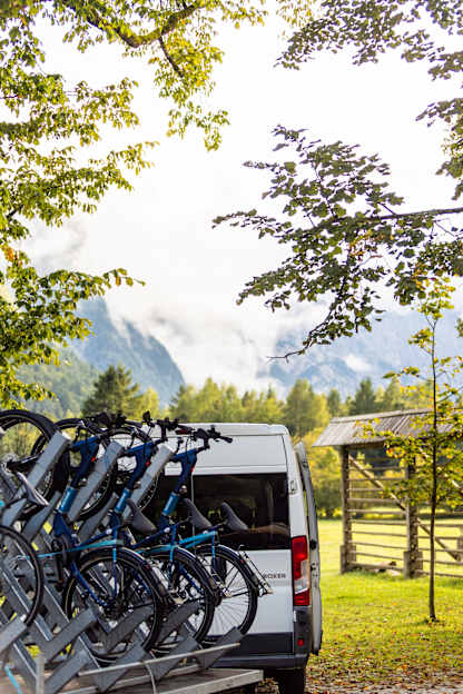 A VBT support van with bikes on a trailer sits beneath leafy trees, backed by misty mountains and a sunlit meadow.