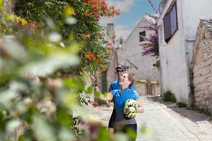Two cyclists pause in a sunny stone alley, admiring bright flowering vines and enjoying a cheerful moment together.