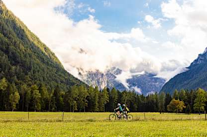 ChatGPT said:  Two cyclists ride through a wide green valley framed by forested mountains and drifting clouds on a bright, scenic day.