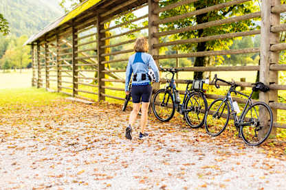 A cyclist walks toward parked bikes beside a rustic wooden shelter, surrounded by autumn leaves and soft mountain light.