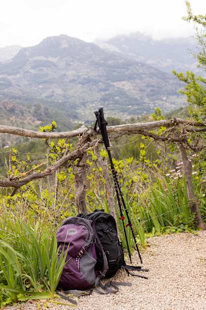 Two backpacks and hiking poles rest against a rustic wooden fence overlooking a misty mountain valley filled with spring greenery.