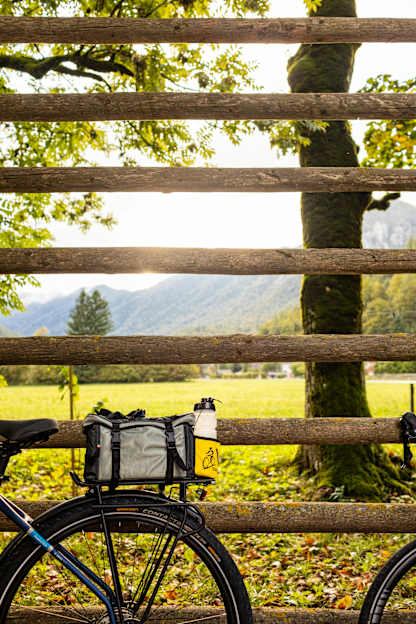 A rustic wooden fence frames a scenic countryside landscape, with a bicycle in the foreground and a lush, green field in the background.