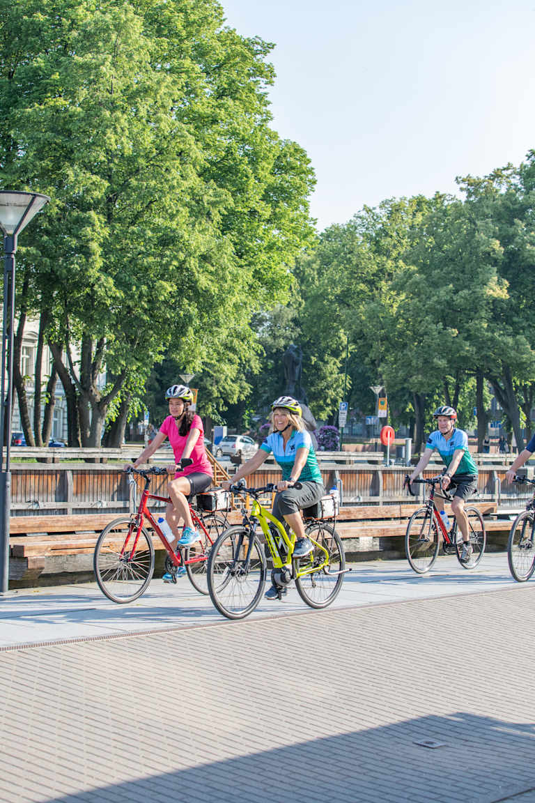 A group of cyclists riding on a paved path surrounded by lush green trees in a park-like setting.