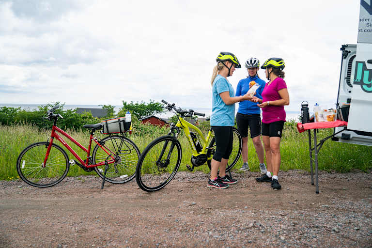Three cyclists in colorful cycling gear standing next to their bicycles in a grassy outdoor setting with a cloudy sky in the background.