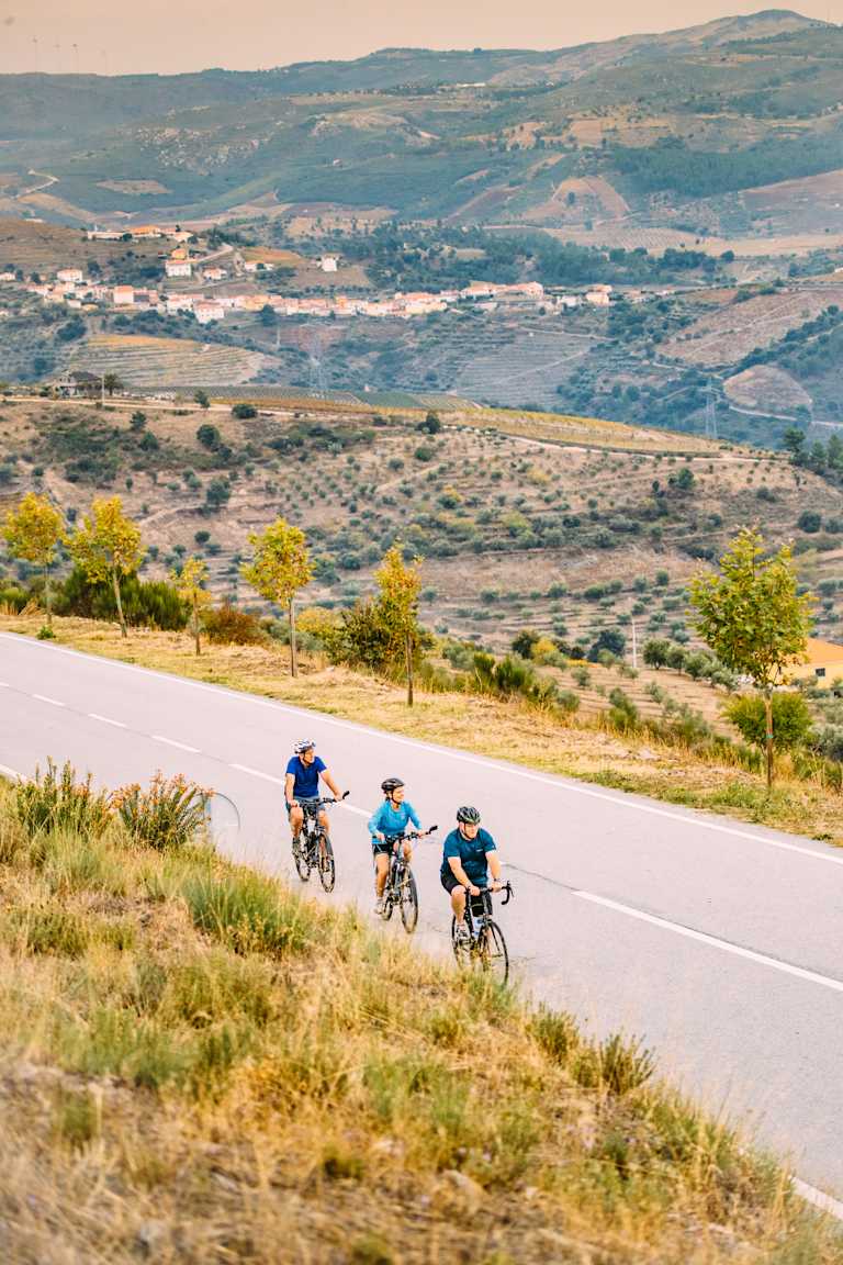 A group of cyclists riding on a winding road through a scenic landscape with rolling hills, distant villages, and a hazy blue sky in the background.