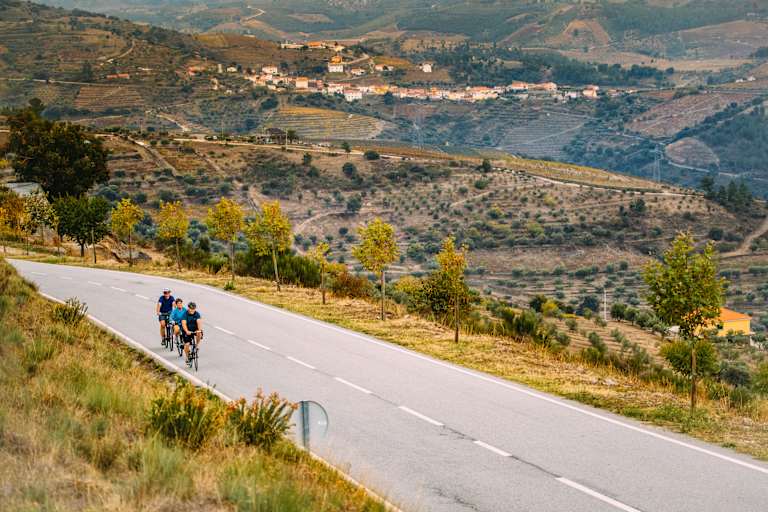 The image depicts a scenic countryside landscape with a winding road in the foreground, where two cyclists are riding, surrounded by rolling hills, olive groves, and a small village nestled in the distance.