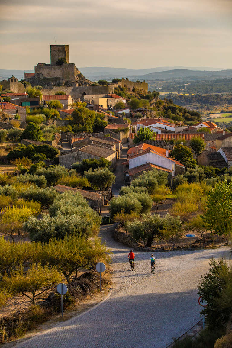 A picturesque village nestled in a hilly landscape, with a medieval castle overlooking the scene and people cycling along a winding road in the foreground.