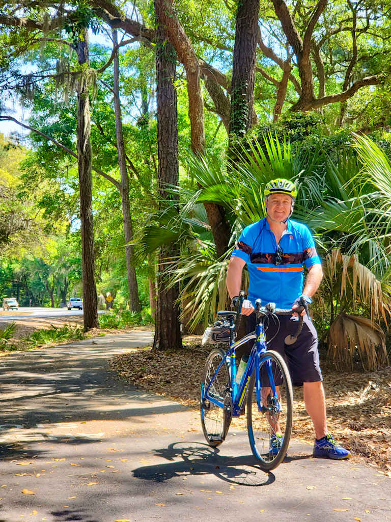 A person in cycling gear stands with a bicycle in a lush, green forest setting with tall trees and palm fronds in the background.