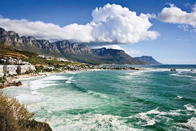 A stunning coastal landscape with towering mountains, a vibrant turquoise ocean, and fluffy white clouds in the bright blue sky.
