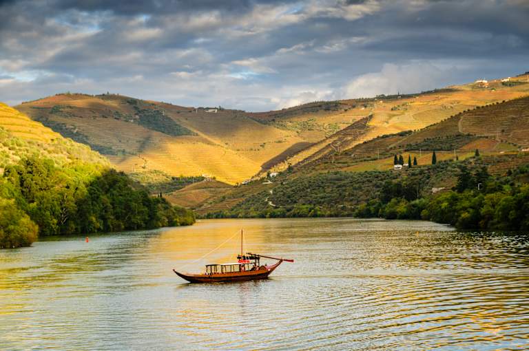 A serene river flows through a picturesque landscape of rolling hills, dotted with trees and a traditional boat sailing across the calm waters, set against a dramatic cloudy sky.