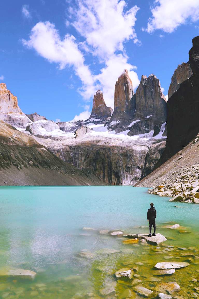 A person stands in the shallow, turquoise waters of a glacial lake, surrounded by towering, snow-capped mountains against a vibrant blue sky with wispy clouds.