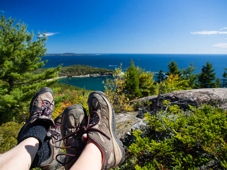 The image shows a pair of hiking boots on a rocky outcrop overlooking a scenic coastal landscape with a deep blue ocean, lush green vegetation, and a clear blue sky.
