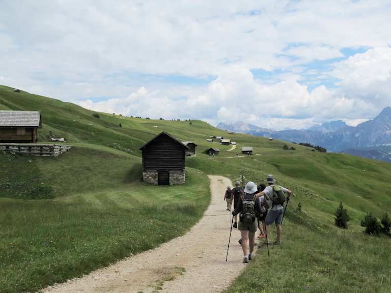 A dirt path winds through a lush, green alpine landscape, with wooden cabins and hikers with backpacks visible in the distance against a backdrop of towering mountains.