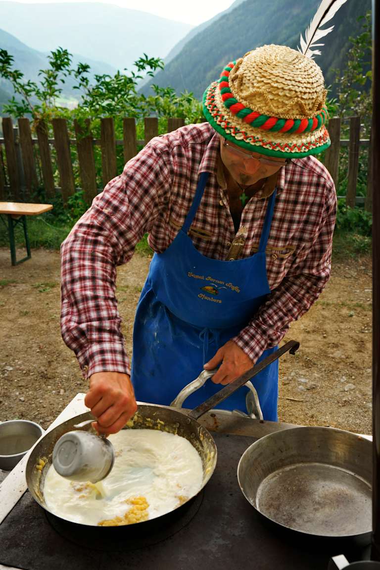 A person wearing a straw hat and plaid shirt is cooking food on a pan outdoors, with a wooden fence and mountains in the background.
