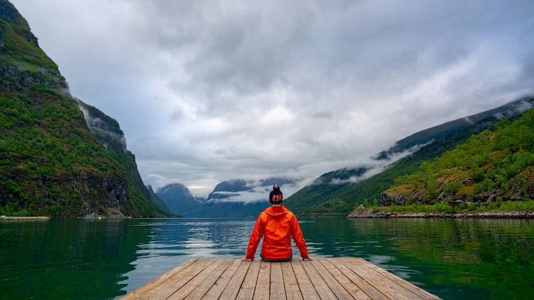 A person in a bright red jacket stands on a wooden dock, surrounded by towering mountains and a serene lake in the background.