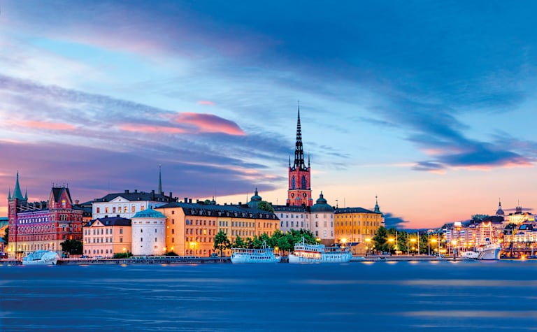 A picturesque cityscape with colorful buildings, spires, and a body of water reflecting the vibrant sunset sky.