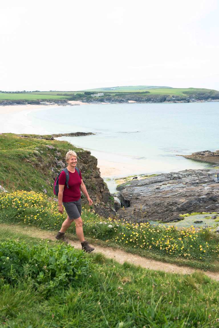 A person in a red shirt and shorts is walking along a grassy path overlooking a scenic coastal landscape with a body of water and distant hills.