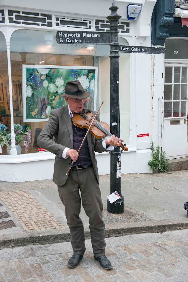 A man in casual attire is playing a violin on the street in front of a museum and art gallery, with a colorful mural visible in the background.