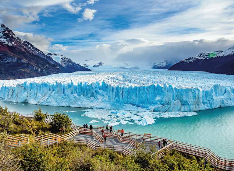 A majestic glacier with towering ice formations rises from a turquoise lake, surrounded by rugged snow-capped mountains and a dramatic cloudy sky, with a wooden walkway leading to the viewpoint in the foreground.
