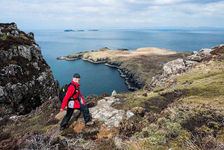 A person in a red jacket stands on a rocky cliff overlooking a vast, serene body of water with distant islands in the background.