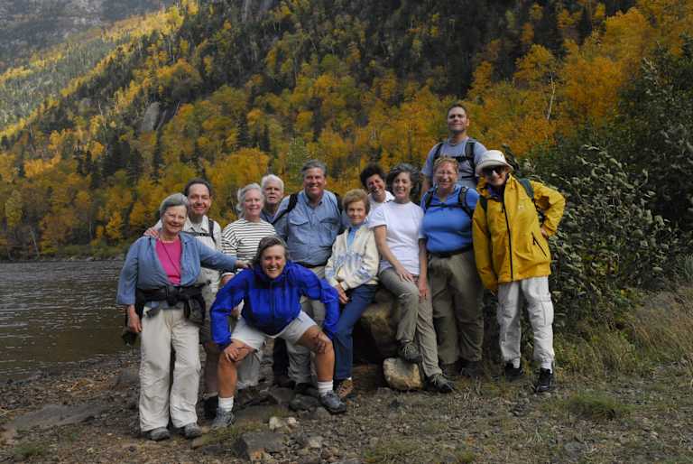 A group of people, dressed in various outdoor attire, stand together in a scenic autumn landscape with a river in the foreground and a forest of colorful trees in the background.