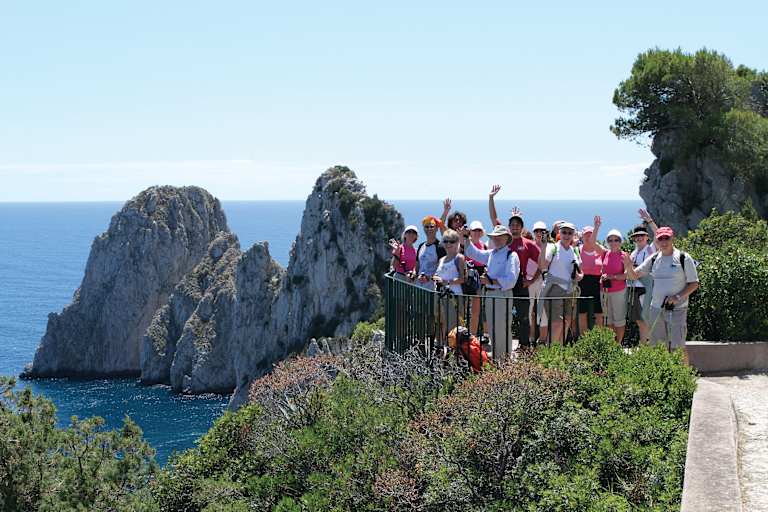 A group of people standing on a viewing platform overlooking a scenic coastal landscape with rocky cliffs and the deep blue ocean in the background.