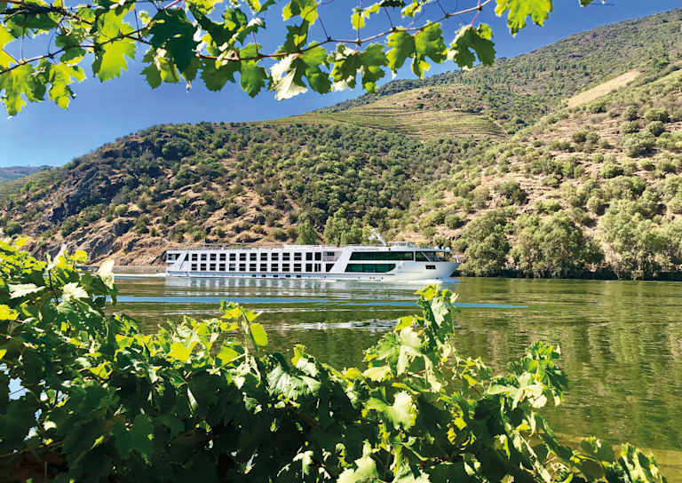 A riverboat cruises along a scenic river surrounded by lush, green foliage and rolling hills in the background.