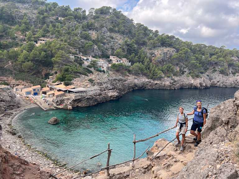 A scenic coastal landscape with a turquoise bay surrounded by rocky cliffs and lush vegetation, with two people walking on a wooden bridge in the foreground.