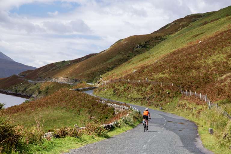 A cyclist rides along a winding road surrounded by lush, rolling hills and a cloudy sky in the background.