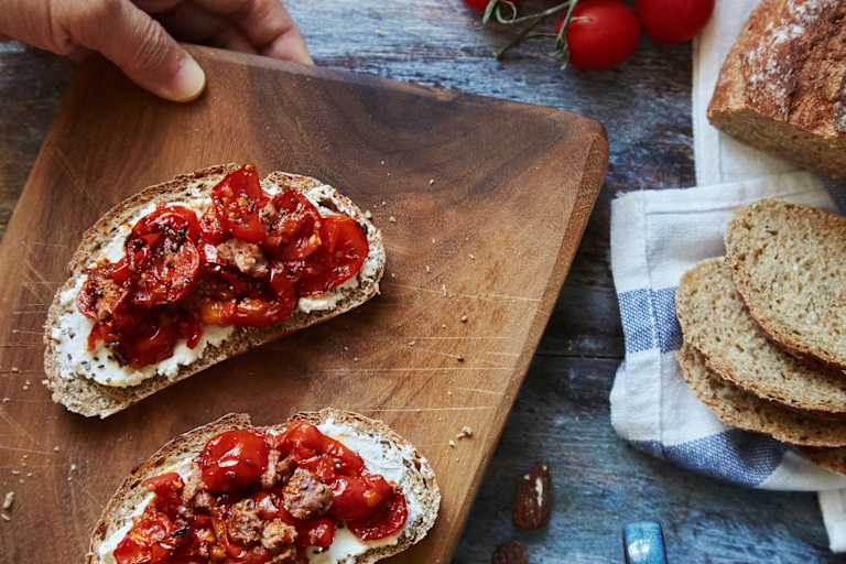 A wooden cutting board with slices of toasted bread topped with a vibrant red tomato-based spread, accompanied by fresh tomatoes and a blue and white striped cloth in the background.