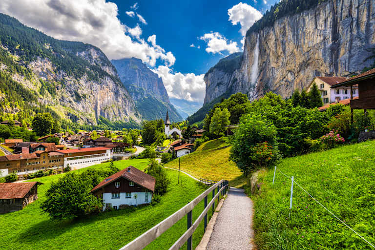 A picturesque alpine village nestled in a lush green valley, surrounded by towering mountains and dramatic cliffs under a vibrant blue sky with fluffy white clouds.