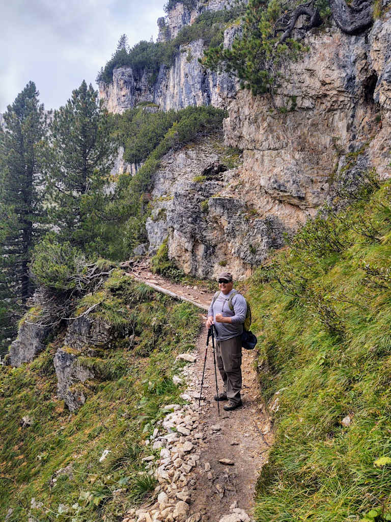 A person is walking along a rocky, winding trail surrounded by lush greenery and towering cliffs in the background.
