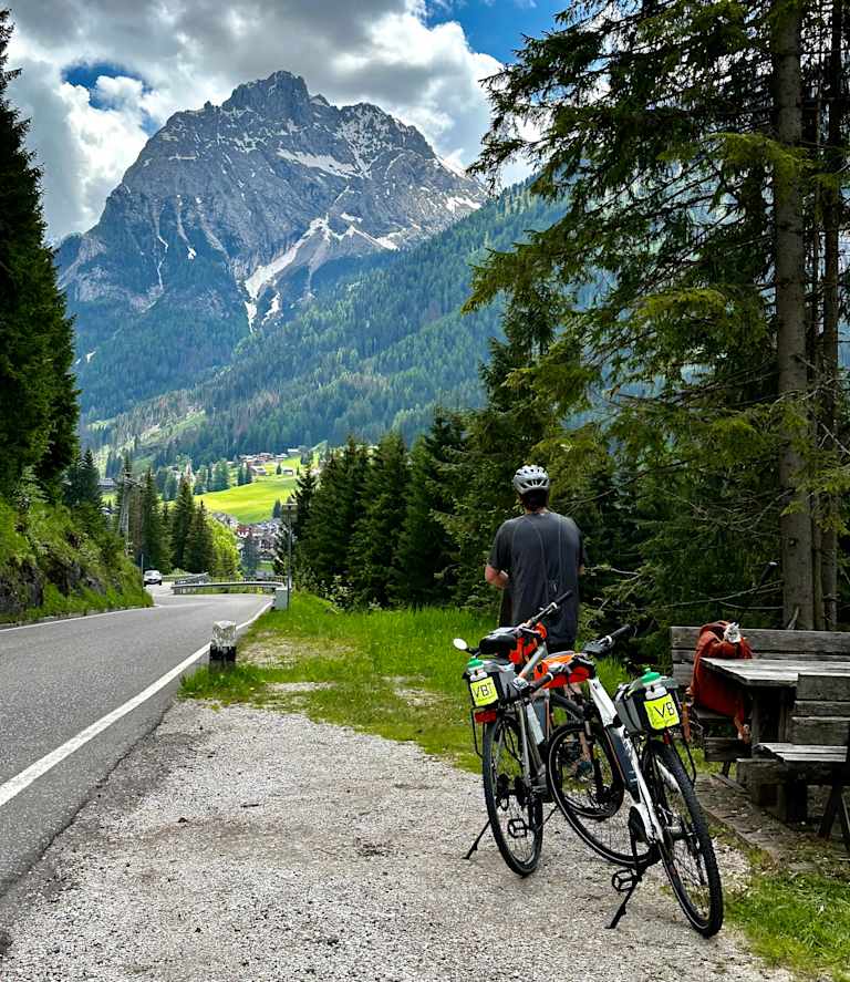 A cyclist stands on a road surrounded by lush green forests and towering snow-capped mountains in the distance.
