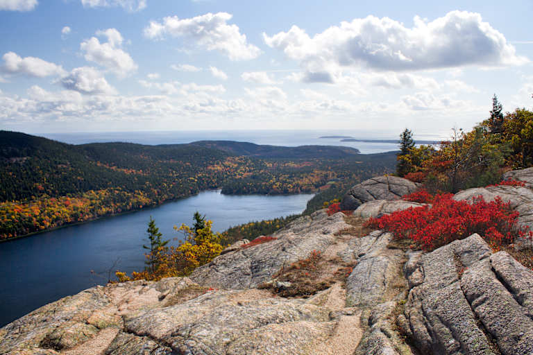 A scenic landscape with a rocky foreground overlooking a vast lake surrounded by vibrant autumn foliage and a cloudy sky in the background.