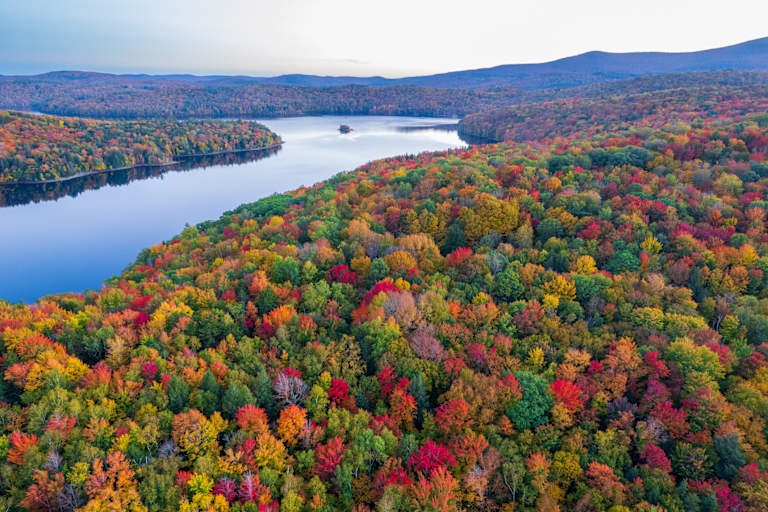 A breathtaking aerial view of a vibrant autumn landscape, with a serene lake surrounded by a lush, colorful forest in the foreground, and rolling hills in the distance.