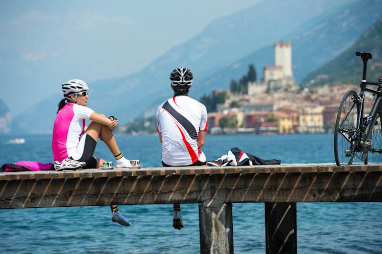 Two cyclists in cycling gear sitting on a wooden dock, with a scenic lakeside town in the background.