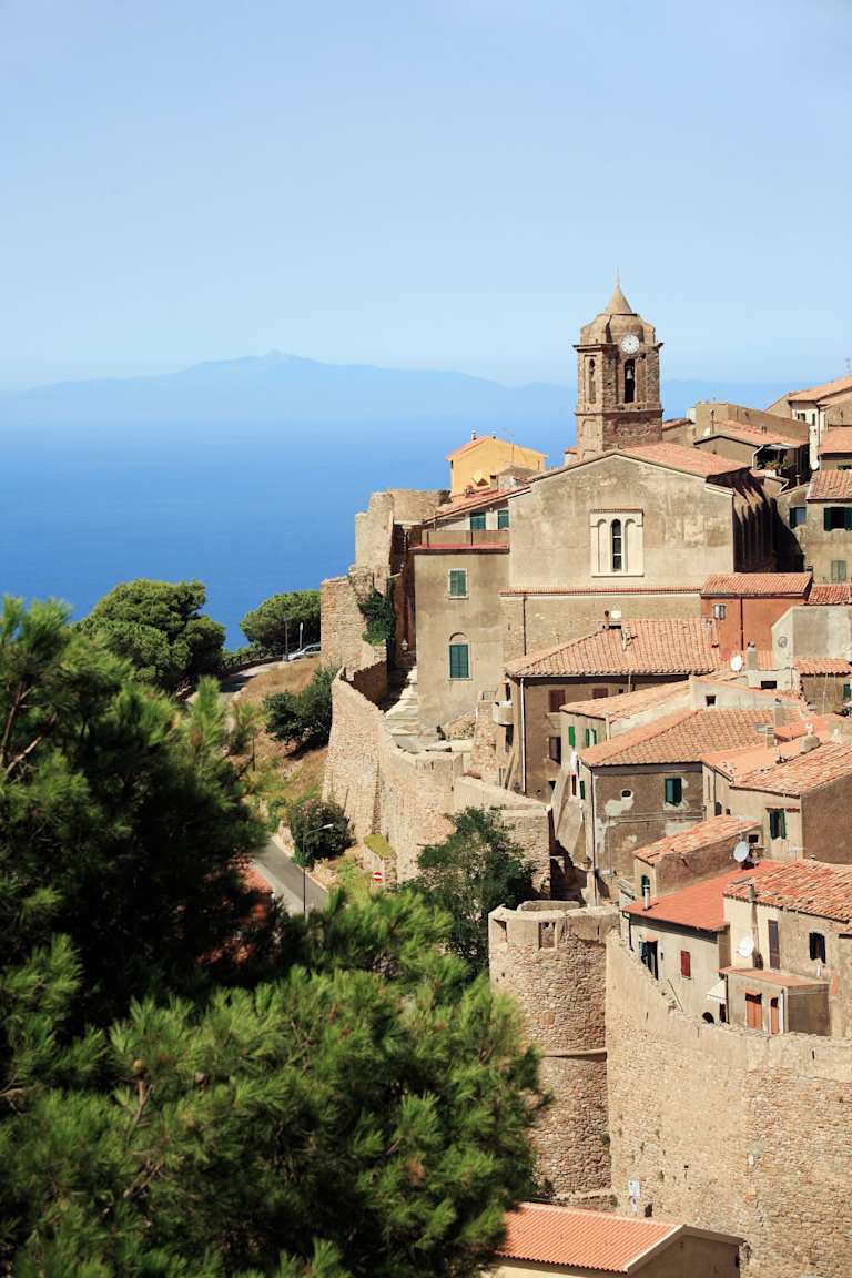 A picturesque Mediterranean village nestled on a hillside, with terracotta-roofed buildings and a bell tower overlooking the vast, blue expanse of the sea in the distance.