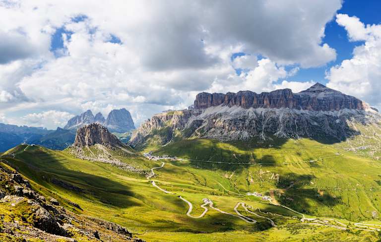 Majestic snow-capped mountains rise against a cloudy sky, with a winding road leading through the lush, grassy foreground.