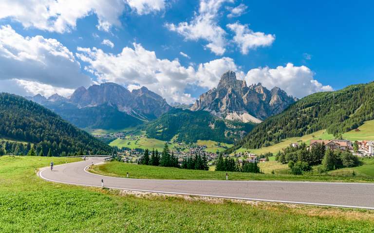 A winding road leads through a lush, green meadow surrounded by towering, snow-capped mountains against a backdrop of a vibrant blue sky with fluffy white clouds.
