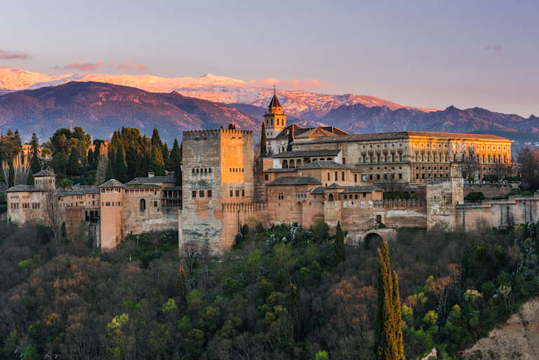 A picturesque medieval castle complex nestled among lush greenery, with majestic snow-capped mountains in the background, bathed in the warm glow of the setting sun.