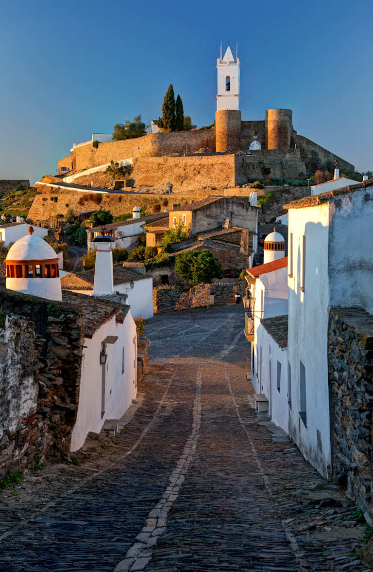 A picturesque hillside town with a historic castle or fortress overlooking a winding path leading through the quaint, whitewashed buildings below.