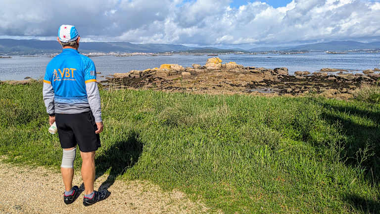 A person wearing a blue shirt and shorts stands in a grassy field overlooking a rocky coastline with mountains in the distance under a cloudy sky.