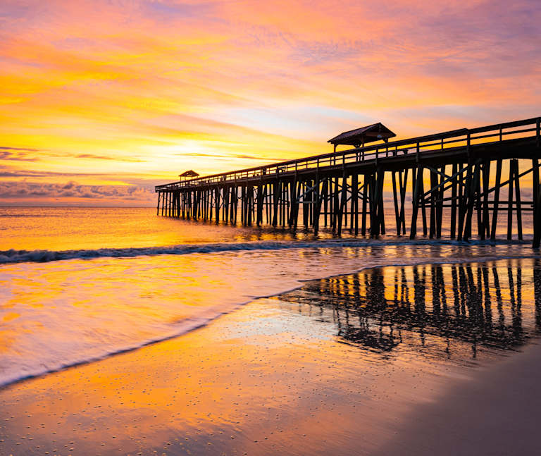 A wooden pier extends into a tranquil ocean, reflecting the vibrant orange and yellow hues of the stunning sunset in the background.