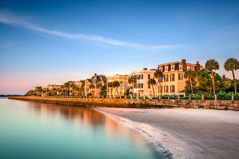 A picturesque coastal town with colorful historic buildings lining the beach, reflected in the calm, turquoise waters of the ocean under a bright blue sky.