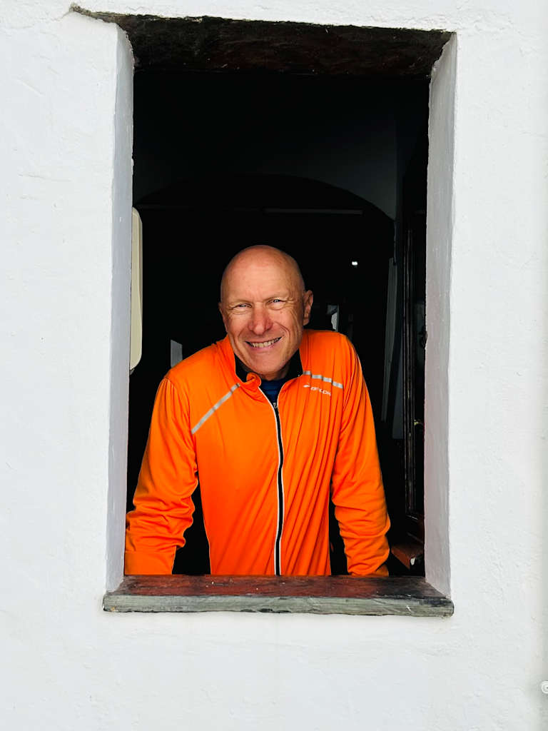 A smiling man in an orange jacket stands in a doorway against a dark background.