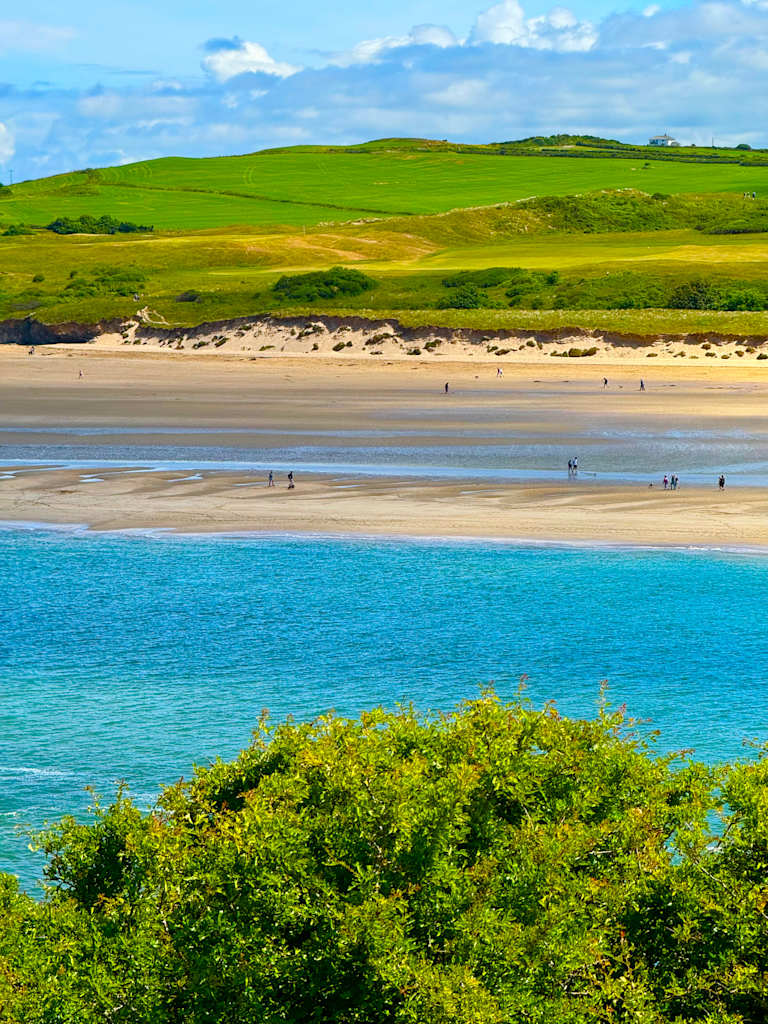 A serene coastal landscape with a sandy beach, lush green hills, and a vibrant blue ocean under a cloudy sky.