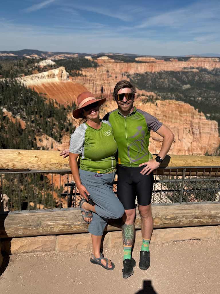Two people, a man and a woman, are standing on a viewing platform overlooking a vast, rugged landscape with a dramatic canyon in the background.