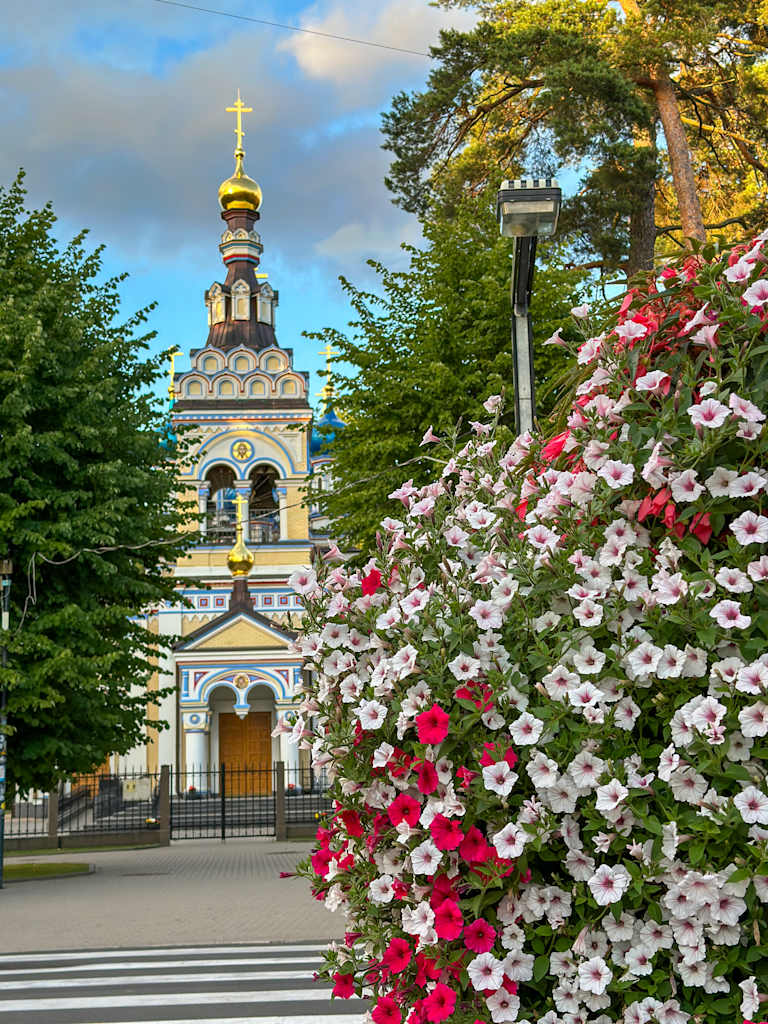 A colorful and vibrant scene featuring a Russian Orthodox church with a golden onion-shaped dome, surrounded by lush greenery and a profusion of blooming flowers in the foreground.