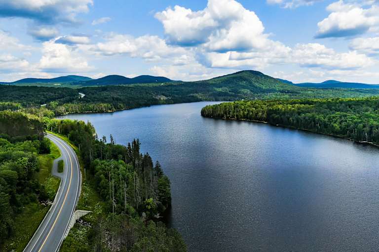 A scenic landscape with a winding road, a serene lake surrounded by lush green forests, and rolling hills in the background under a cloudy blue sky.